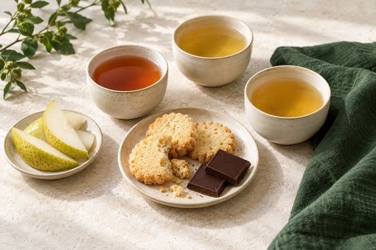 Ivory tea cups with shortbread, dark chocolate and pear slices on a pale stone table with forest-green linen.
